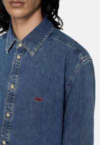 Camicia in denim blu scuro con colletto a punta, chiusura con bottoni e logo ricamato rosso sul lato sinistro del petto. Texture liscia.