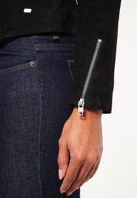 Schwarze Wildlederjacke mit einem silbernen Reißverschluss und einer strukturierten Oberfläche, kombiniert mit dunklen Jeans aus Denim, die über kontrastierende Nähte und ein Knopf-Detail verfügen.