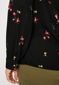 Sort langærmet bluse med rød og gul blomstermønster, knaplukket manchet, båret med olivengrønne bukser, hånd afslappet ved siden.