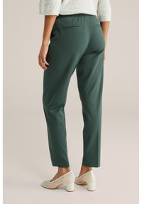 Nő sötétzöld egyenes szárú nadrágban és krémszínű alacsony blokksarkú cipőben, semleges háttér előtt állva.