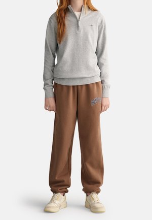 Person wearing light gray quarter-zip sweatshirt, brown sweatpants with "GANT USA" text, and beige sneakers, standing against white background.