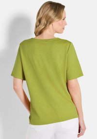 Grünes strukturiertes T-Shirt mit kurzen Ärmeln und rundem Ausschnitt, von hinten gezeigt. Kombiniert mit weißen Hosen.