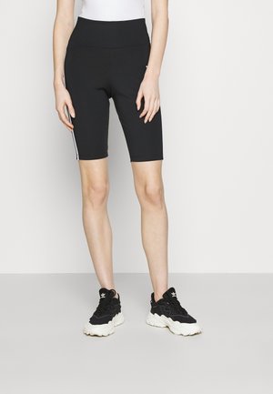 Person iført sorte knælange tætsiddende shorts med hvide sidelister og sorte og hvide sportssko, stående mod en ensfarvet baggrund.
