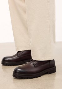 Bocanci din piele maro cu o textură netedă, vârf rotund și talpă robustă. Acei aurii pentru șireturi. Purtați cu pantaloni deschiși la culoare, cu picior drept.