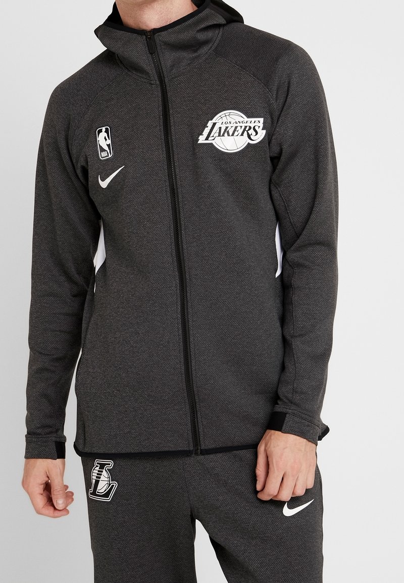 Sort Nike zip-up jakke med Los Angeles Lakers-logo, tekstureret stof og hvide sidestykker; båret med matchende sorte bukser og et Nike-logo.