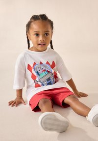 Fehér pamut póló színes grafikával, amely egy kutyát ábrázol a szemetesben, piros rövidnadrággal és fehér sportcipővel párosítva. Világos háttér előtt ülve.