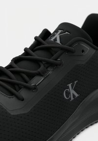Black athletic shoe with a textured mesh upper, flat laces, a shiny accent near the eyelets, and a subtle logo on the side.