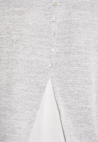 Top de punto gris claro que presenta un patrón texturizado, seis botones blancos en la parte delantera y una abertura en la espalda que revela una capa blanca inferior.