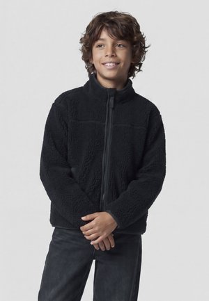 Garçon aux cheveux bouclés souriant, portant une veste polaire noire zippée et un pantalon foncé sur un fond clair uni.