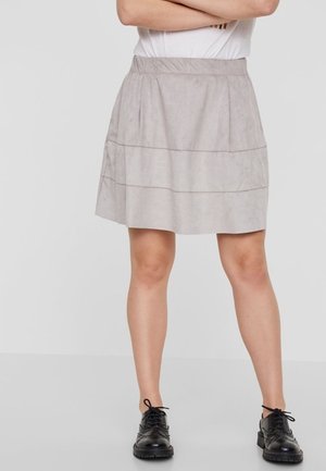 Lichtgrijze, suede-achtige rok met een elastische tailleband; heeft horizontale stiksels en een uitlopende vorm. Zwarte schoenen eronder gedragen.