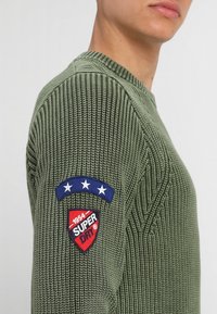 Man in een olijfgroene gebreide trui met een blauwe patch met drie witte sterren en een rode schildpatch met de tekst "1954 SUPER DRY" op de mouw.