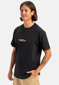 Camiseta de algodón negra con un logo frontal en blanco, diseño de cuello redondo, mangas cortas y un ajuste relajado. Sin patrones ni acentos adicionales.