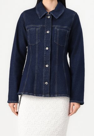 Mujer vistiendo una chaqueta de mezclilla ajustada de color azul oscuro con botones en la parte frontal y dos bolsillos en el pecho, sobre una falda blanca texturizada.