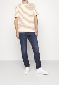 Camiseta de manga corta, cuello redondo, de rayas naranjas y blancas combinada con jeans azul oscuro y zapatillas blancas. Ajuste casual, material de algodón.