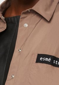 Světle hnědá košile na knoflíky s černým štítkem na kapse, na kterém je nápis "esmé stu", nošena přes černý top.