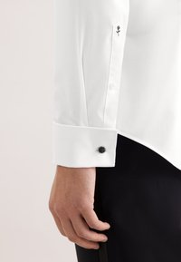 Męska ręka przy mankiecie białej koszuli z czarnym spinką do mankietu, małym czarnym haftem róży na rękawie oraz czarne spodnie.