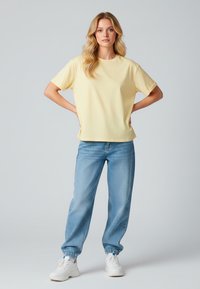 T-shirt di cotone gialla con vestibilità rilassata e maniche corte, abbinata a pantaloni in denim blu e sneakers bianche. Design semplice, senza motivi.