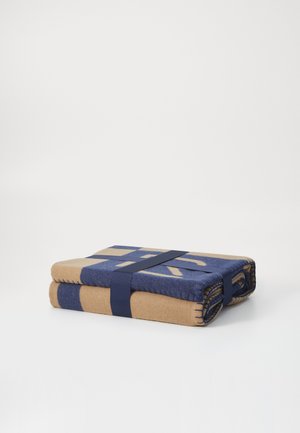 Folded blanket in tan and navy blue, featuring a geometric pattern. Secured with a navy strap. The fabric appears soft and textured.