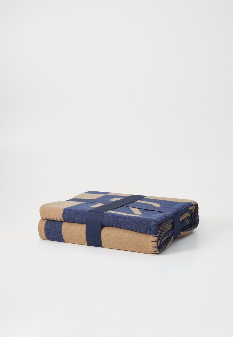 Coperta piegata in beige e blu navy, con un motivo geometrico. Fissata con una cinghia blu navy. Il tessuto sembra morbido e testurizzato.