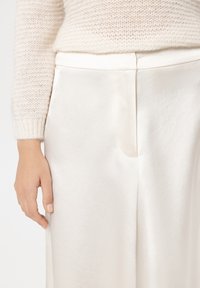 Weiße Satin-Hosen mit glatter Textur, versehen mit einer seitlichen Nahtdetail und einem weit geschnittenen Bein, kombiniert mit einem leichten Strickpullover.