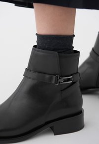 Sorte lær ankle boots med en rem og sølvspenne, teksturert overdel og lav blokkhæl. Ullsokk som stikker ut.
