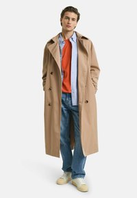 Beige trenchcoat med dobbeltrækket design over en rød sweater og en lys blå skjorte. Sammen med blå jeans og hvide sneakers.