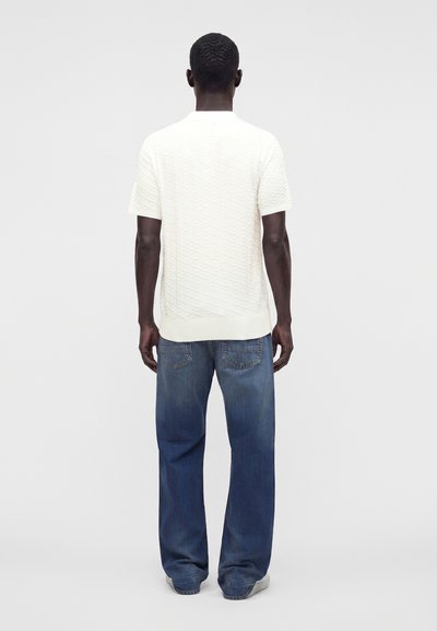 Homme debout, de dos, portant une chemise blanche texturée à manches courtes et un jean bleu ample, sur un fond clair uni.