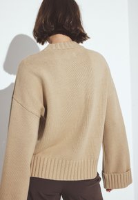 Person med kort brunt hår som bär en överdimensionerad beige stickad tröja med breda ärmar och mörkbruna byxor, vänd bort från kameran.