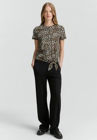 Korte mouwtop met luipaardprint in beige en zwart, voorzien van een knoop aan de zoom. Gecombineerd met zwarte wijde broekspijpen en strakke zwarte schoenen.