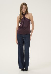 Bordeaux satijnen haltertop met een gedrapeerde voorkant, gecombineerd met brede, donkere denim jeans. Zwarte spitse schoenen. Neutrale achtergrond.