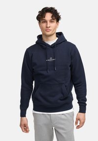 Sweat à capuche bleu marine en tissu doux, avec une poche avant et un cordon de serrage. Le logo "KRONSTADT" est affiché sur la poitrine.