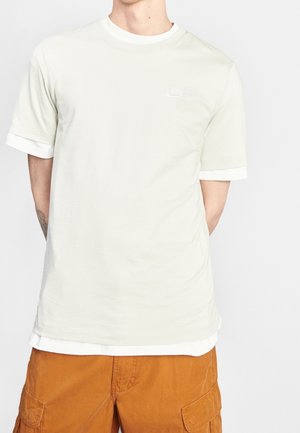 Person trägt ein hellbeiges Nike-Kurzarmshirt über einem weißen Shirt, kombiniert mit orangefarbenen Cargo-Shorts, die Arme hinter dem Rücken verschränkt.