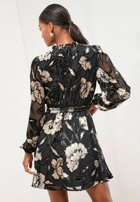 Melnas ziedu kleitas ar zelta ziedu motīviem, garām piedurknēm un savilktām kakla līnijām. Izgatavota no caurspīdīga auduma ar taisnu vidukli.