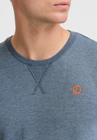 Blå crewneck sweatshirt med tekstureret stof, der har et synligt orange broderet logo på brystet og en ribbenkrave.