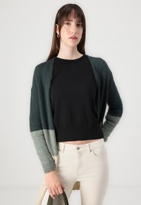 Largo abrigo verde y gris con frente abierto, usado sobre una camiseta negra y jeans de color claro. Textura suave con puños acanalados.