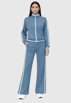 Femme debout portant un survêtement bleu avec des bandes blanches sur les manches et le pantalon, assorti à des baskets blanches, sur un fond gris.