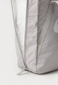 Sac à dos en tissu gris avec une surface texturée, doté d'une ouverture zippée et d'un intérieur blanc. Comprend une poignée latérale et un détail de logo.