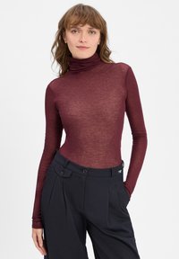 Burgundská dlhá mikina s rolákom vyrobená z priehľadnej látky, spárovaná s čiernymi vysokými nohavicami so strihom na mieru, ktoré majú dva gombíky v páse.
