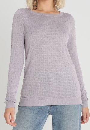 Jasnofioletowy, długi sweter o teksturowanej strukturze z prążkowanymi mankietami i rantem, noszony z jasnoniebieskimi dżinsami przez stojącą osobę.