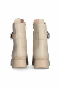 Stivaletti beige alla caviglia con tacco a blocco, cerniera laterale, cinghie decorative e fibbie in metallo, mostrati da dietro su uno sfondo bianco.