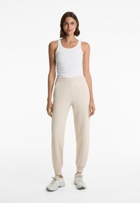 Woman standing wearing a white sleeveless tank top, beige high-waisted jogger pants, and white sneakers on a white background.