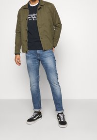 Giacca leggera verde oliva sopra un maglione blu navy, abbinata a jeans denim azzurri e sneakers nere. Stile casual, vestibilità rilassata.