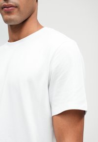 Λευκό βαμβακερό T-shirt με στρογγυλή λαιμόκοψη και κοντά μανίκια, με λεία υφή και μινιμαλιστικό σχεδιασμό, απεικονίζοντας μια κοντινή λήψη της λαιμόκοψης και του μανικιού.