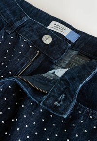 Tamsiai mėlynos džinsinės plačios kojos kelnės su aukštu liemeniu, dekoruotos sidabriniais cablais, metaliniu užtrauktuku ir firminiu žymekliu viduje.