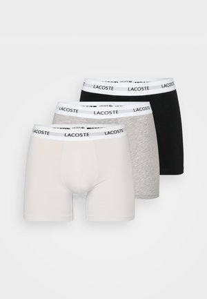 Tři páry pánských boxerek v bílé, šedé a černé barvě. Každý pár má bílý pás s opakovaným logem "Lacoste". Měkký bavlněný materiál.