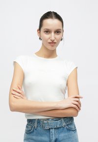 Λευκό μπλουζάκι με κοντά μανίκια και εφαρμοστή γραμμή, με ανάγλυφη λεπτομέρεια σε σχήμα αστεριού. Συνδυάζεται με μπλε τζιν παντελόνι ψηλόμεσο.