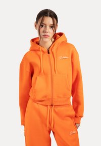 Haut à zip orange en mélange de coton, doté d'une poche avant, de poignets côtelés et d'un logo brodé sur la poitrine. À associer avec un pantalon assorti.