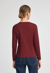 Bordeaux långärmad skjorta gjord av mjukt tyg, med en enkel rund halsringning och en figurnära passform, i kombination med mörkblå jeans.