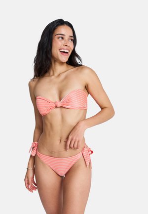 Femme souriante, portant un bikini bandeau rose corail à rayures blanches avec un bas à nouer sur les côtés, sur un fond uni.