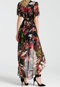 Robe maxi en tissu noir transparent, ornée de motifs floraux et de feuilles vifs. Manches courtes, taille cintrée et ourlet asymétrique. Talons hauts noirs.